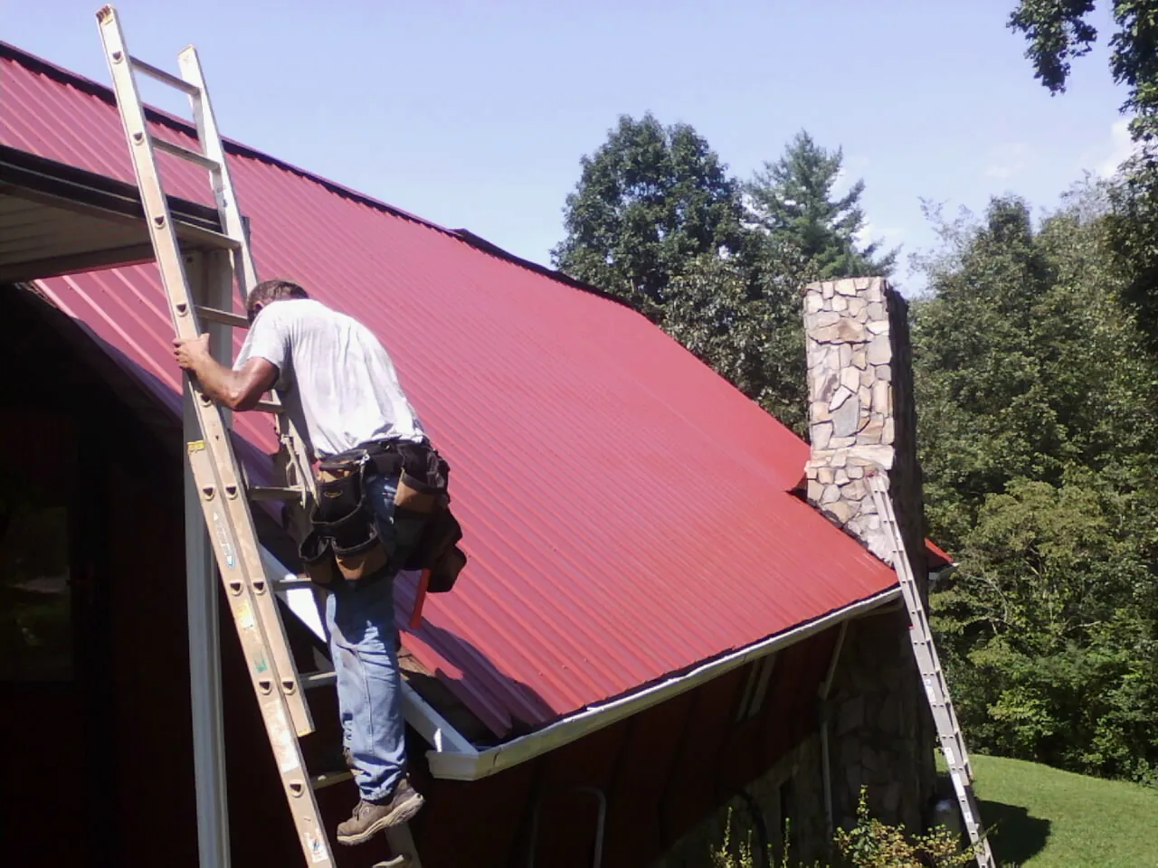 Roofer on a ladder beside a red metal roof and stone chimney — Soffit & Fascia Repair by Roofing Brothers in Mohawk